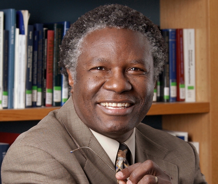 Calestous Juma holds his glasses while sitting in front of a bookshelf